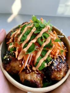 Handheld view of a ceramic bowl filled with Banh Mi Meatballs, jasmine rice, and cilantro sprigs.