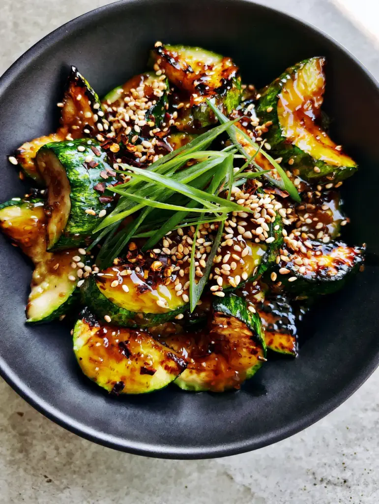 A minimalist matte black bowl filled with seared zucchini topped with sesame seeds and chili flakes. (Zucchini stir fry recipe)