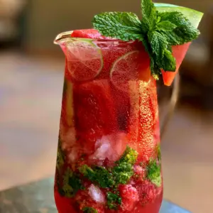 Close up of a condensation-covered pitcher showing layers of crushed ice, mint leaves, and watermelon mojito liquid. (watermelon mojito)