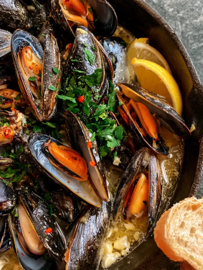 Close up of steamed blue mussels with orange meat and fresh flat-leaf parsley in a cast iron pan.