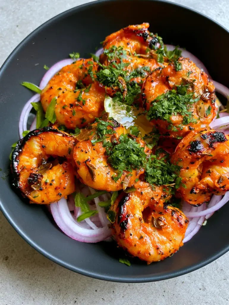 Charred tandoori prawns served on a bed of red onions and green chili slivers in a black bowl.